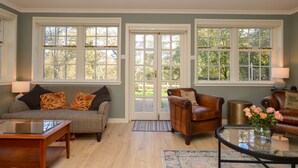 A spacious living room with a sofa, armchairs, and a coffee table. There are large windows with a view of trees and a door leading outside.