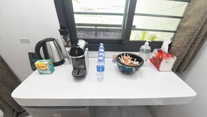 Una pequeña cocina con una encimera blanca, una tetera, una cafetera, una botella de agua, un cuenco con aperitivos y una caja de pañuelos.