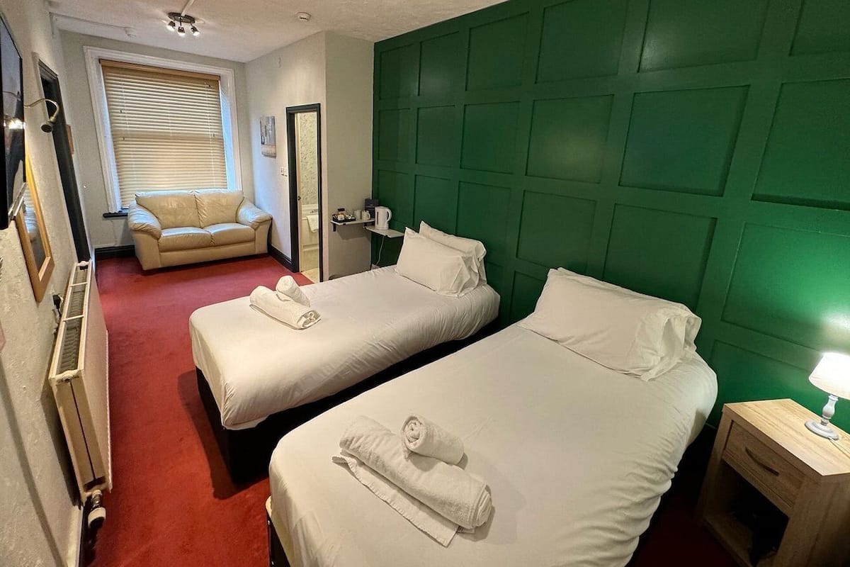 A hotel room with two beds, a green headboard, a small bedside table with a lamp, a window with blinds, and a chair.