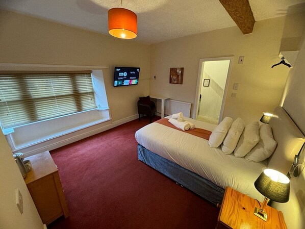 A bedroom with a bed, a TV, a window with blinds, a mirror, and a lamp.