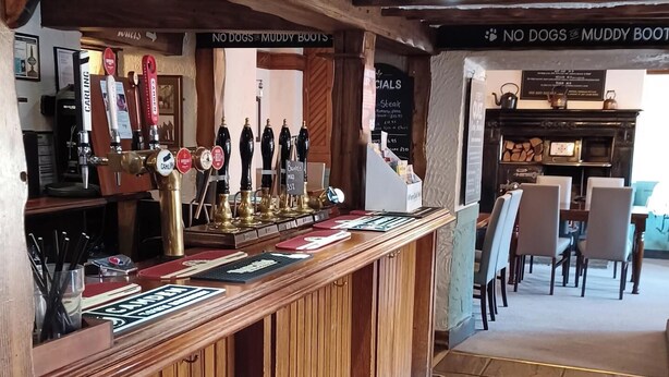 A bar area with wooden beams, a counter with taps, and a menu board. - George Hotel Hayfield (High Peak)