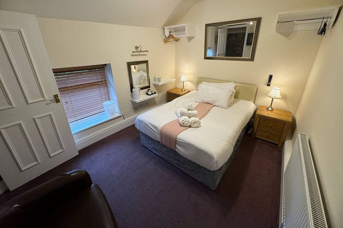 A neatly arranged hotel room with a bed, bedside lamps, a window with blinds, a mirror, and a radiator.