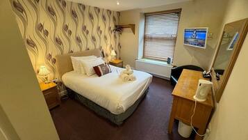 A hotel room with a bed, a desk, a chair, a radiator, a window with blinds, a lamp, and a TV screen displaying a woman named Laura.