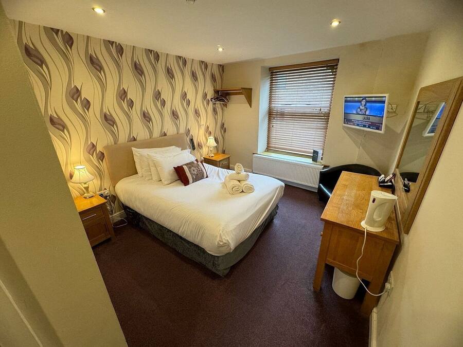 A hotel room with a bed, a desk, a chair, a radiator, a window with blinds, a lamp, and a TV screen displaying a woman named Laura.