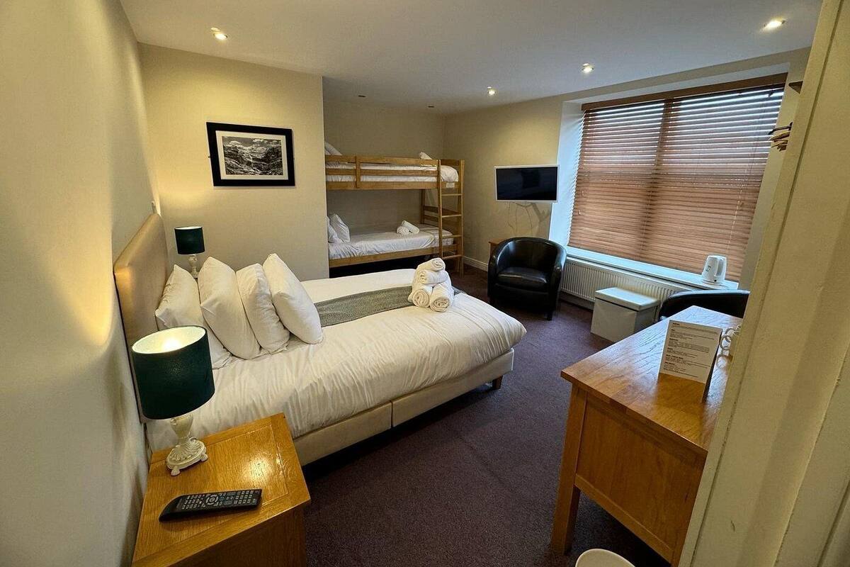 A hotel room with a single bed, a desk with a chair, a TV, and a bunk bed in the background.