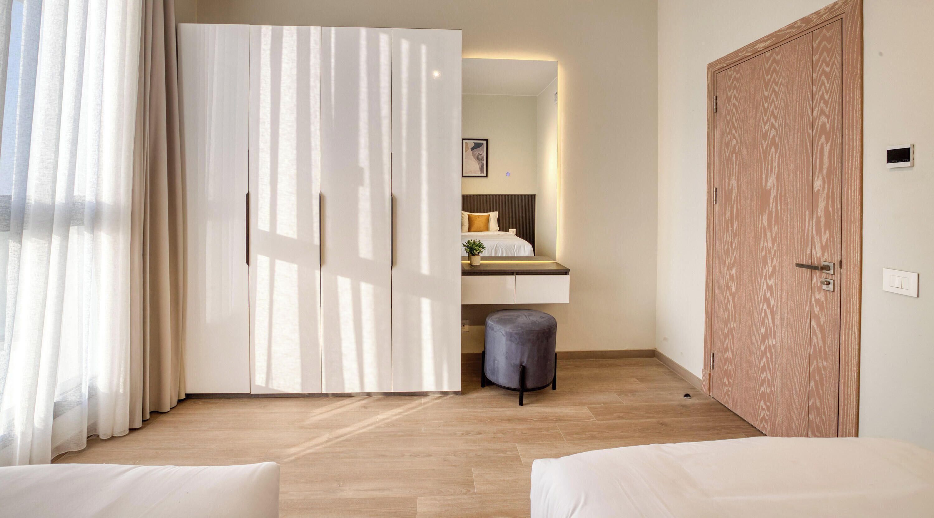 A hotel room with a large wardrobe, a bedside table with a lamp, and a small stool.