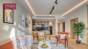 A modern living room with a sofa, armchair, and coffee table. There is a kitchen area in the background with a dining table and chairs. - Prime Select Mall of Arabia (6th of October City)