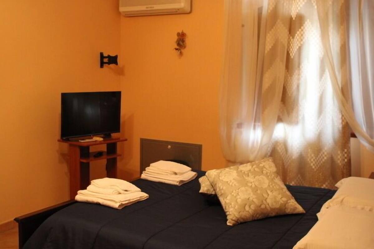 Una habitación de hotel con una cama, un televisor sobre un mueble y una ventana con cortinas traslúcidas.