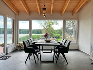 A dining area with a table set for six, large windows offering a view of a lake and greenery, and a wooden ceiling.