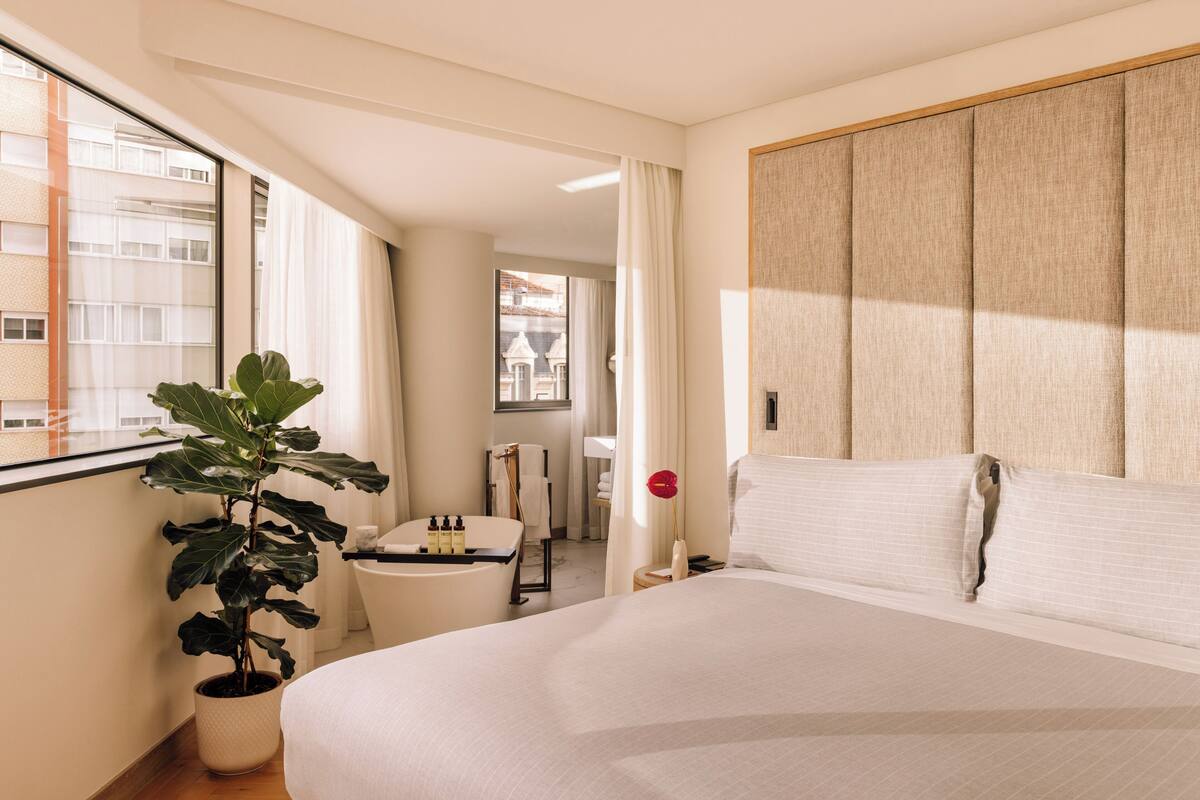 Een hotelkamer met een groot bed, een kamerplant, een klein tafeltje met toiletartikelen en uitzicht op de stad.