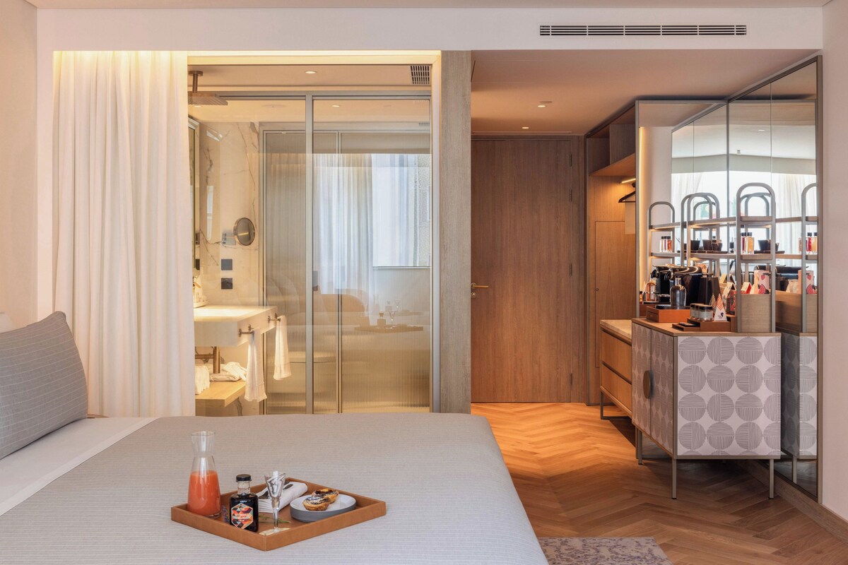 Une chambre d'hôtel moderne comprenant un lit, une table de chevet avec un plateau de petit-déjeuner, une armoire et une salle de bain visible à travers une porte en verre.