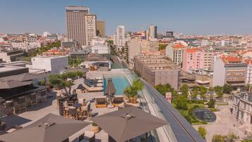 A rooftop pool area with lounge chairs and umbrellas, surrounded by urban buildings and a clear sky.