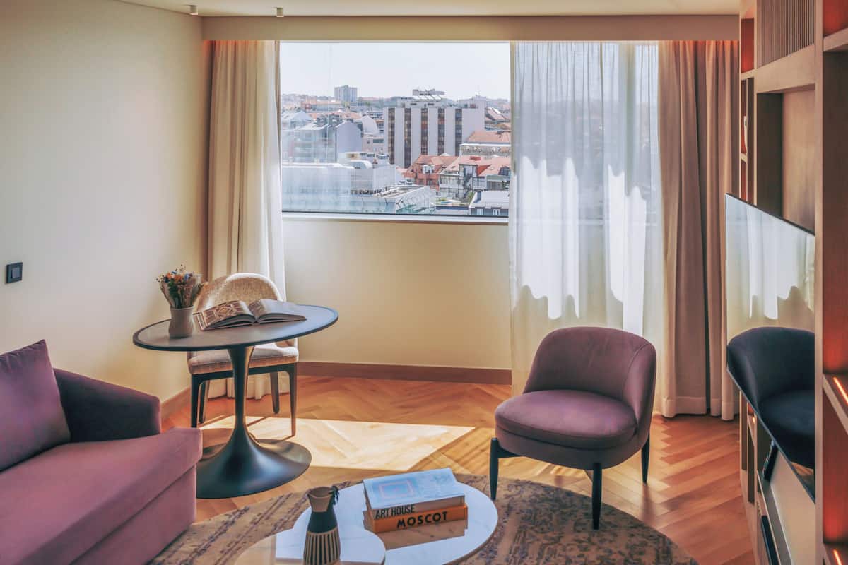 Een moderne hotelkamer met een bank, fauteuil, een klein tafeltje met een boek en een groot raam met uitzicht op de stad.