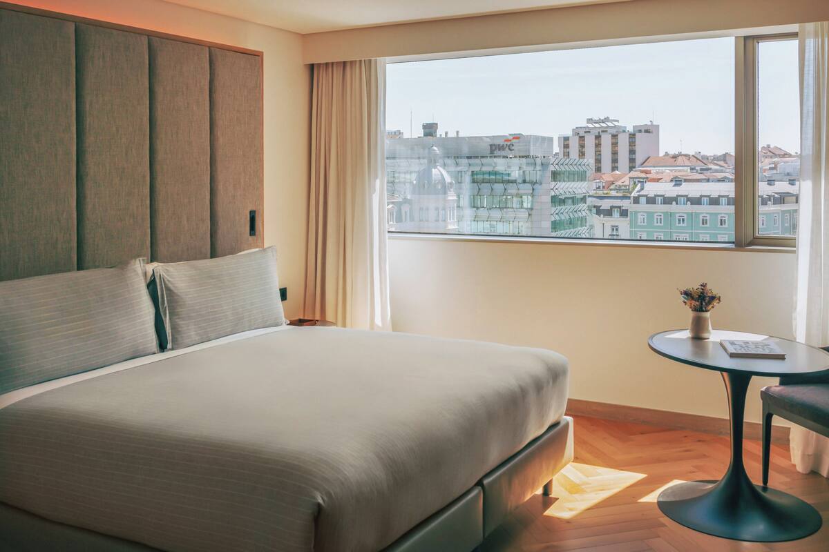 Hotelkamer met een groot bed, een klein rond tafeltje en uitzicht op gebouwen door een raam.