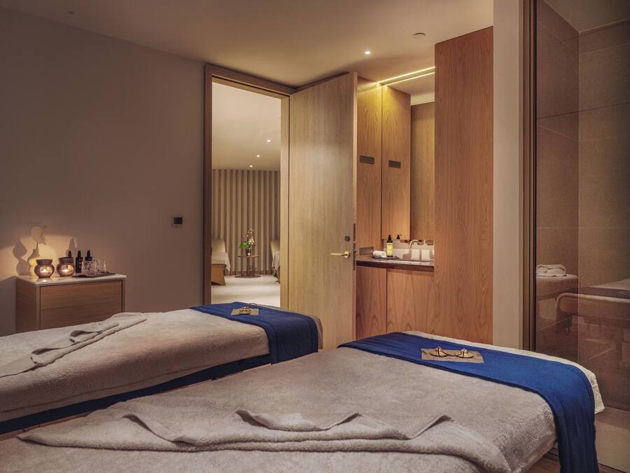 A hotel room with two beds, a bedside table with a lamp, a mirror, and a bathroom visible through an open door.