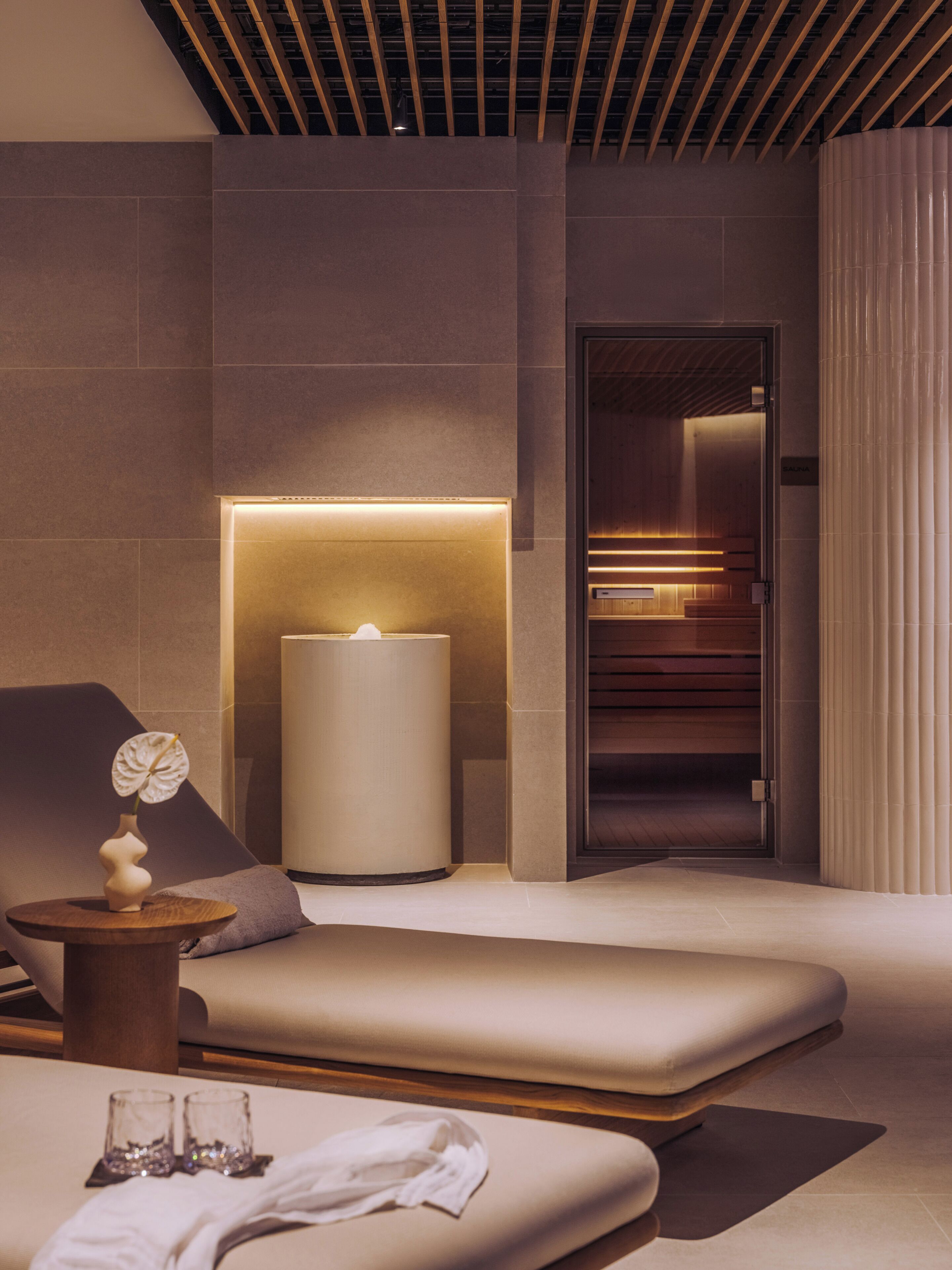 A modern spa room with a massage table, a wall-mounted massage chair, a small table with a vase and glasses, and a door leading to a sauna area.