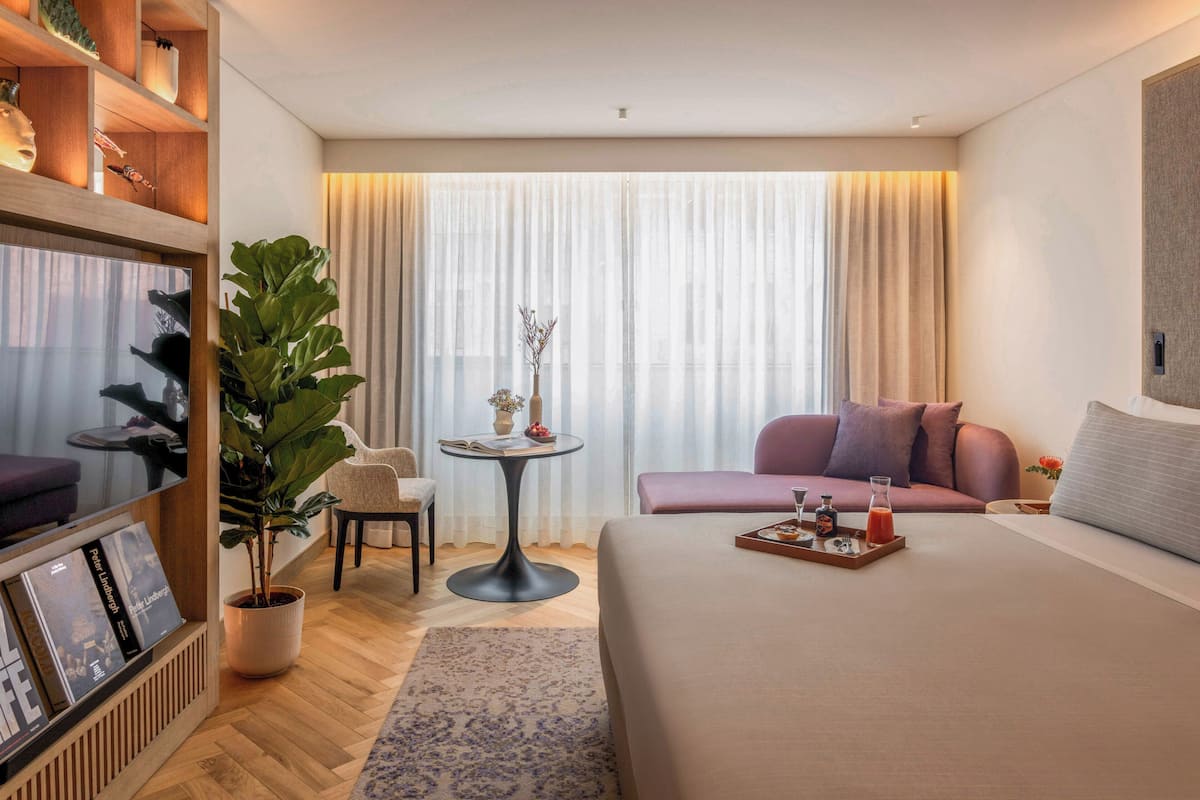 Een moderne hotelkamer met een groot bed, een zithoek met een bank en stoelen, een klein rond tafeltje met een vaas en boeken, en een grote kamerplant.