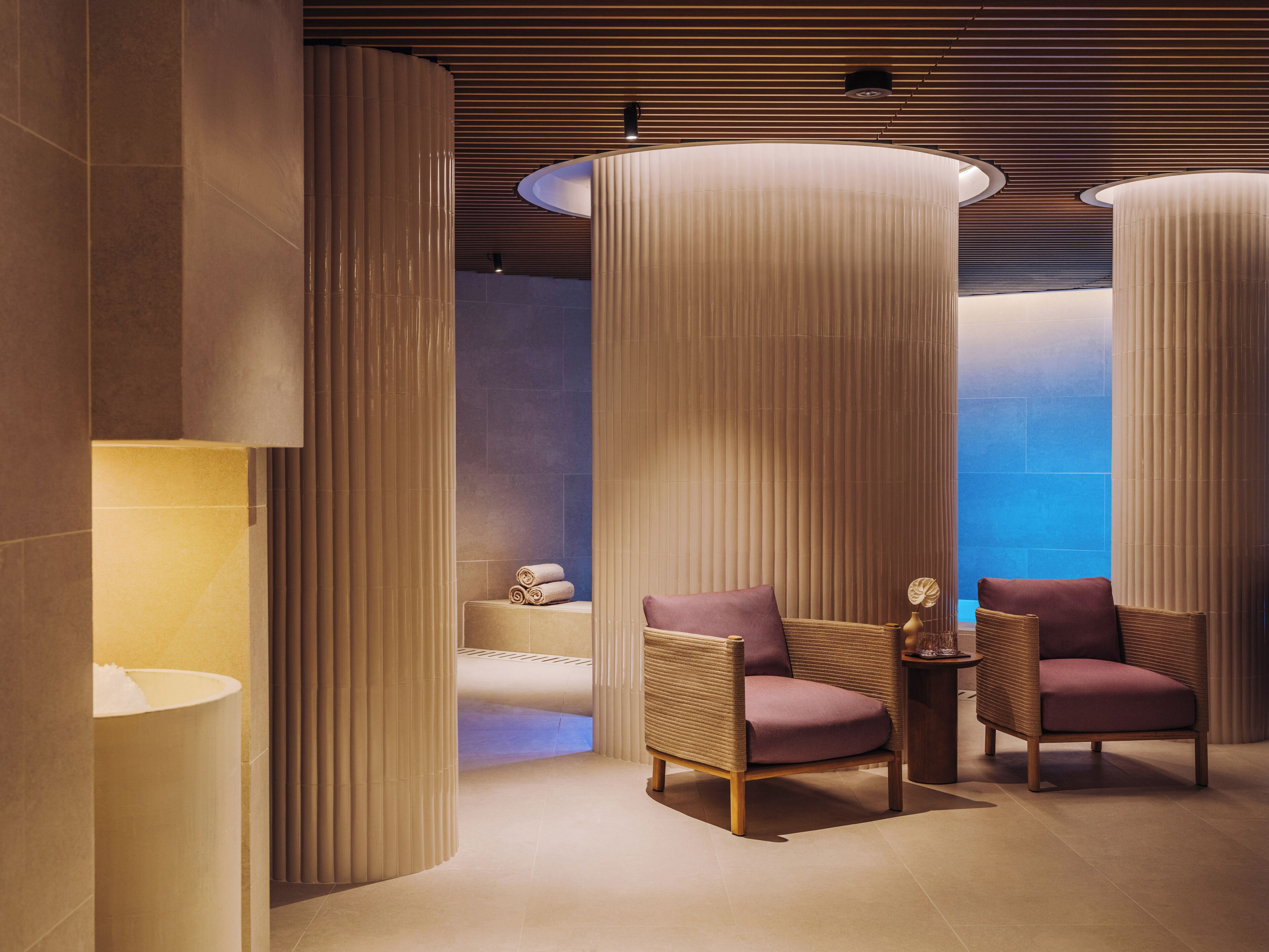 Une salle de spa moderne, dotée de deux fauteuils en osier, d’une table de massage et de hautes colonnes cylindriques.