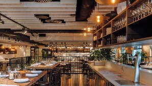 Un restaurant avec une longue table en bois dressée pour le repas, de nombreux tabourets de bar et des étagères remplies de verres et de bouteilles.