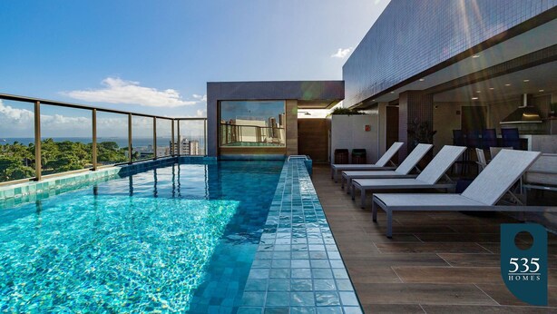 Ein Infinity-Pool auf dem Dach mit Liegestühlen, einer Glasbrüstung und Blick auf die Stadtlandschaft.