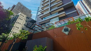 A modern high-rise building with a glass facade, balconies, and a wooden fence with greenery. - 2 Bedroom Apartment with Sea View Rooftop (Salvador)