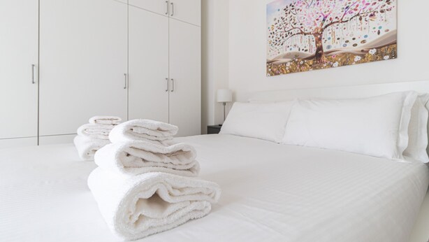 A neatly made bed with white linens and a stack of folded towels on the floor. - Italianway - Serra 16 (Milan)
