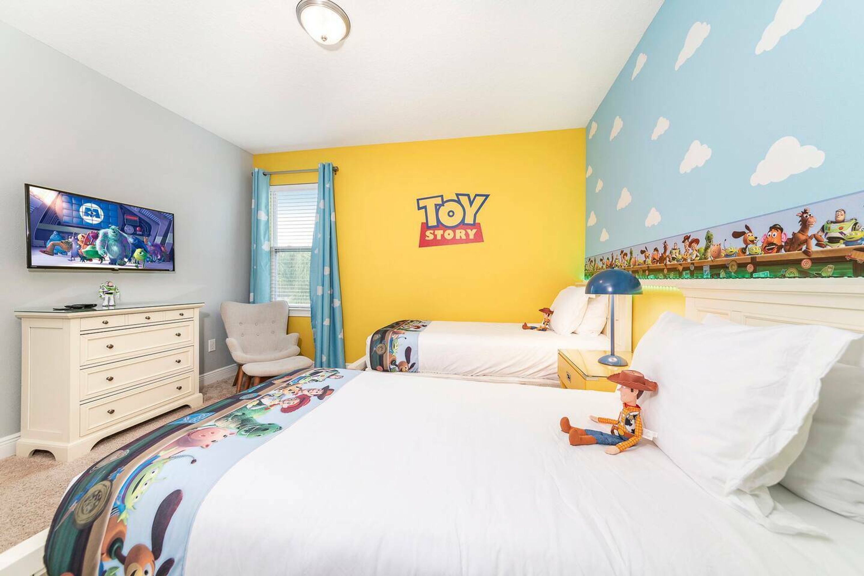 A bedroom with a yellow wall, a 'Toy Story' mural, a TV, a dresser, two beds, and a window with blue curtains.