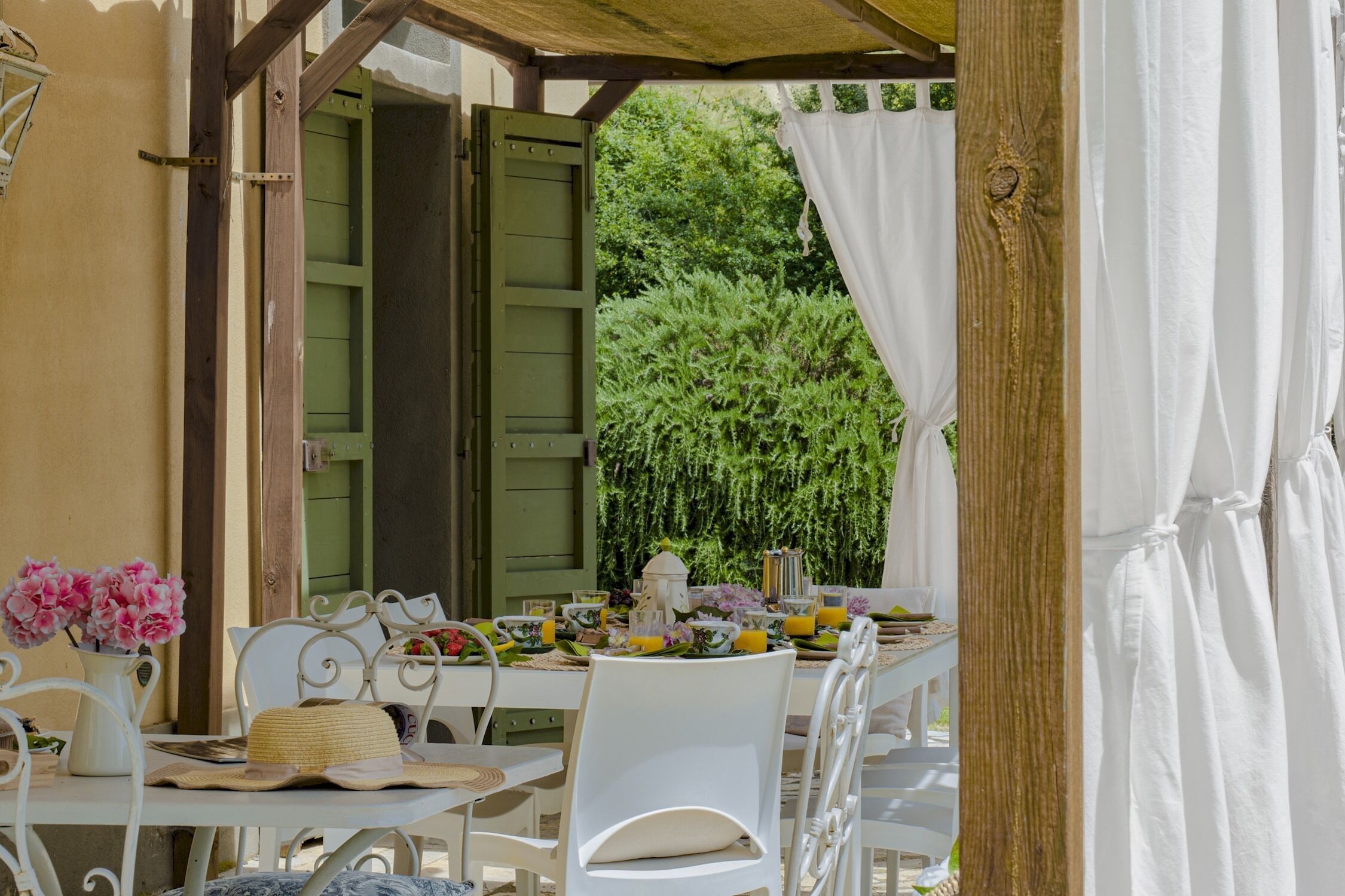 An outdoor dining area with a table set for two, surrounded by white chairs, a wicker basket, and a pitcher of flowers.