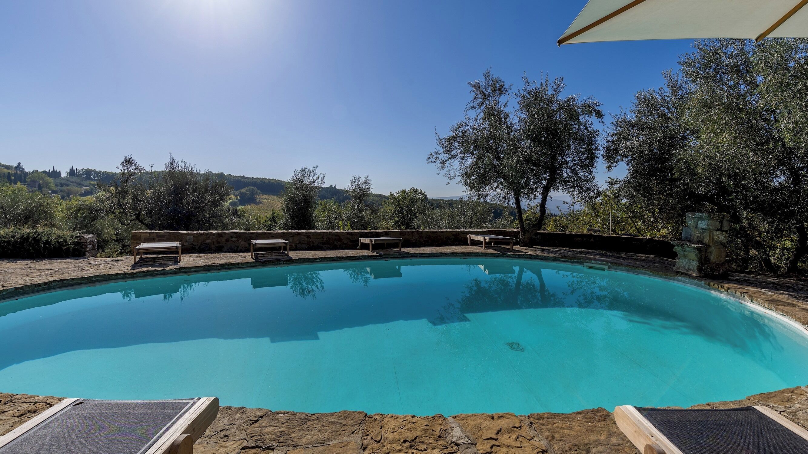 A clear blue swimming pool with lounge chairs and a stone wall, surrounded by trees and a view of hills.