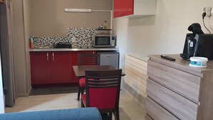 Une cuisine compacte équipée d’un four à micro-ondes, d’une machine à café et d’un petit réfrigérateur. On y trouve une table à manger avec des chaises, ainsi qu’un meuble en bois doté de tiroirs.