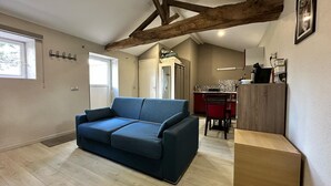 Un salon moderne avec un canapé bleu, des poutres en bois au plafond, un coin cuisine avec une chaise rouge et un meuble en bois.