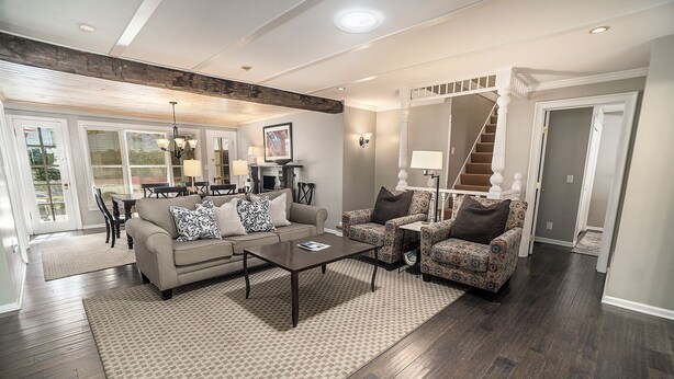 A spacious living room with a large sofa, a patterned armchair, and a coffee table. There is a staircase leading to another level, and a dining area is visible in the background.