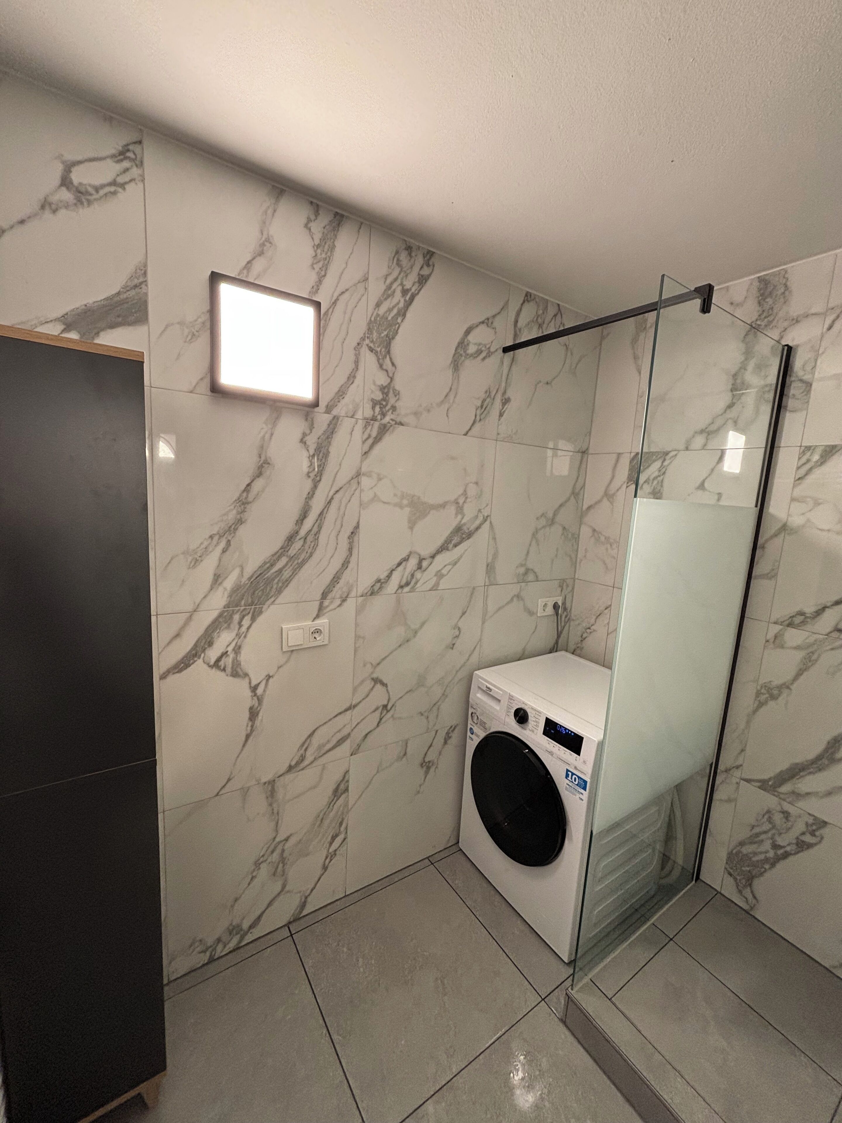 A compact laundry room with a washing machine, a glass shower enclosure, and a marble-patterned wall.