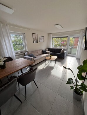A modern living room with a sofa, a dining table, and chairs. There are large windows providing natural light and a view of the outdoors. - Nisay Home - Schorndorf - City Apartment (Schorndorf)
