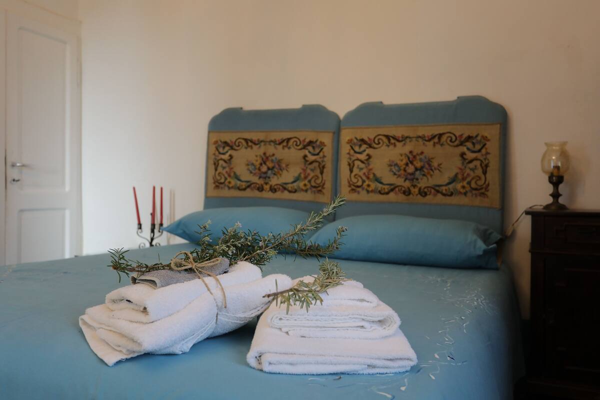 A neatly made bed with folded white towels and a sprig of rosemary, a bedside lamp, and a wooden nightstand.
