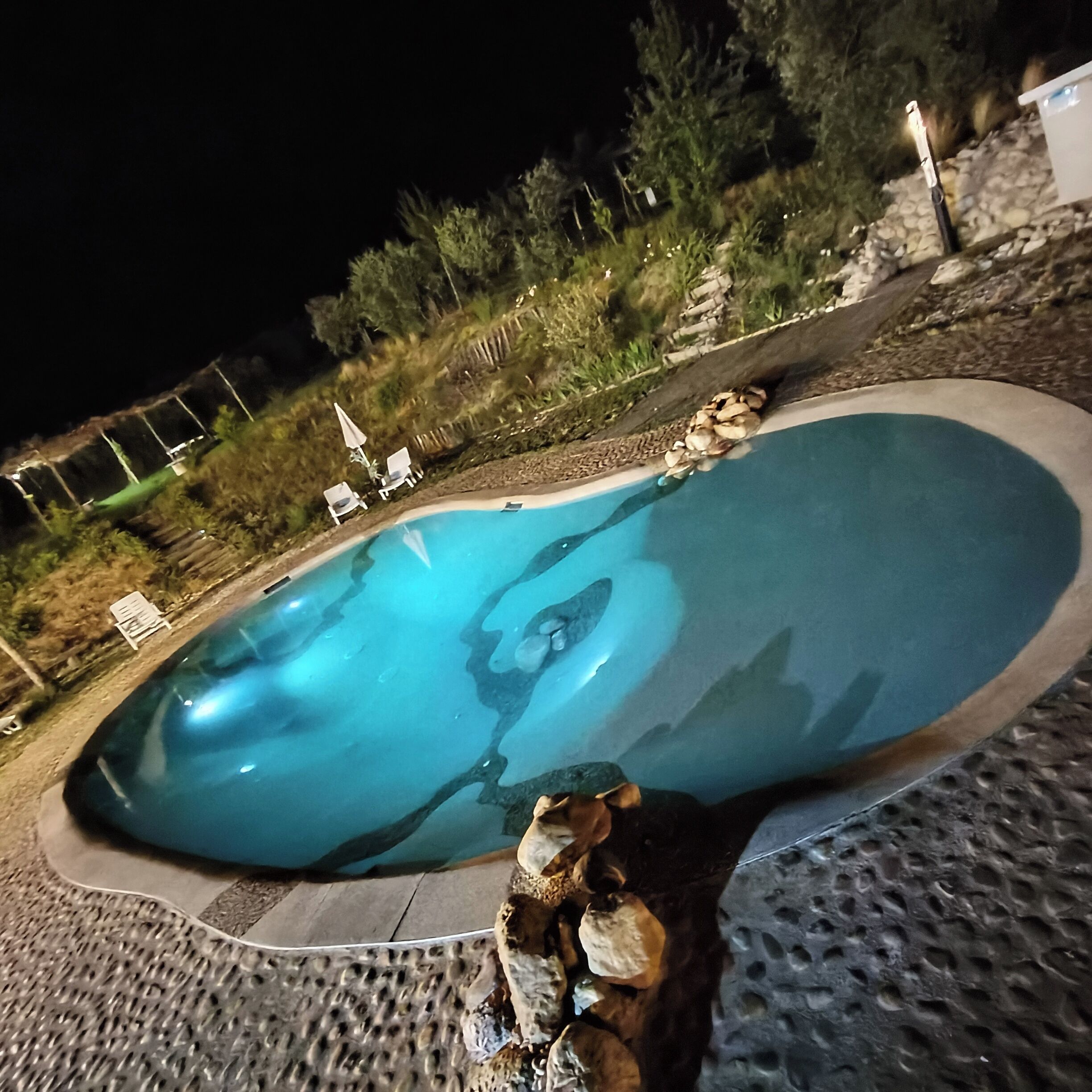 Une piscine bordée de pierres, entourée d’une terrasse avec des chaises longues et un petit jardin.