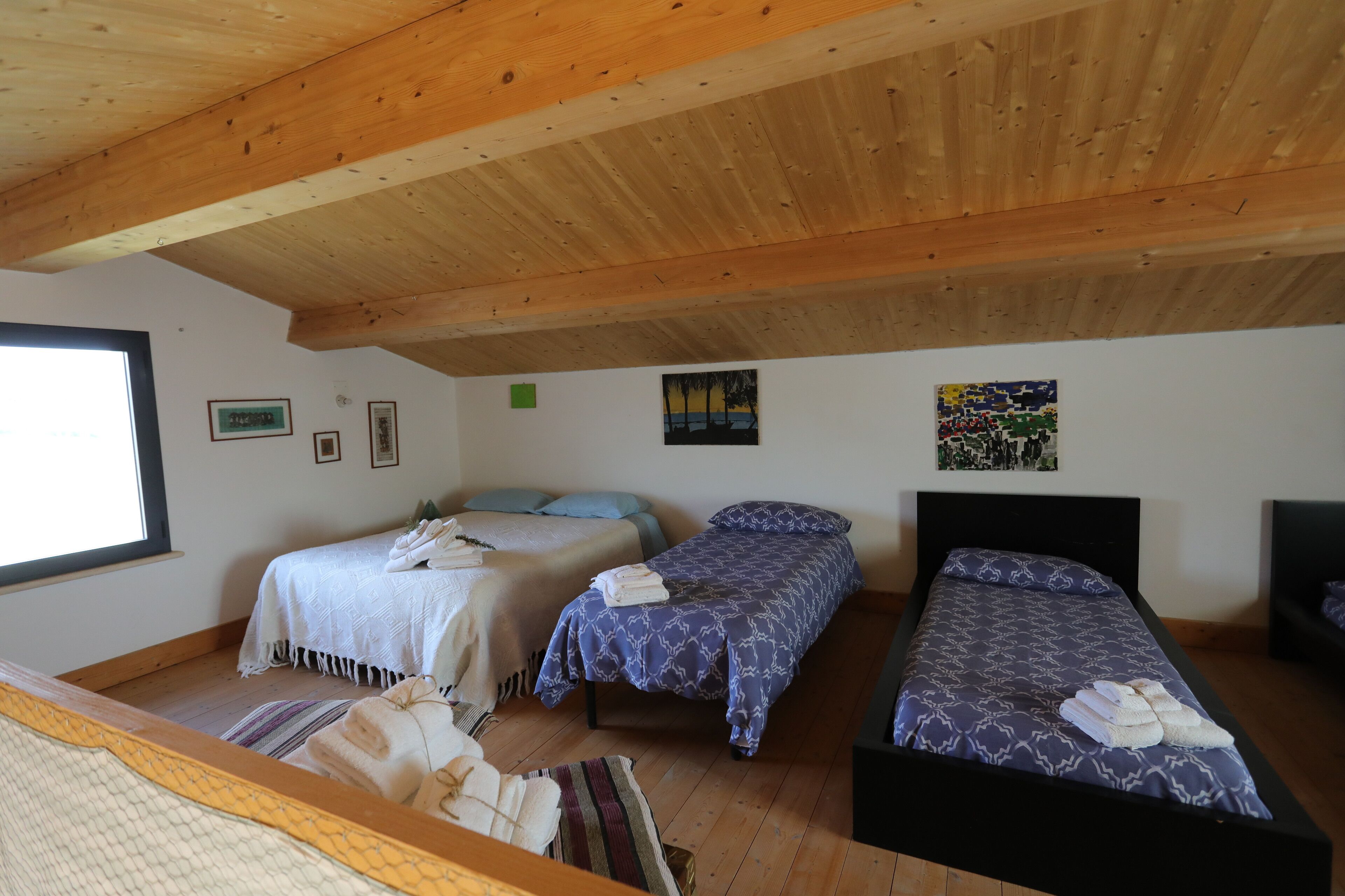 Une chambre avec trois lits, des poutres apparentes en bois au plafond et une grande fenêtre.