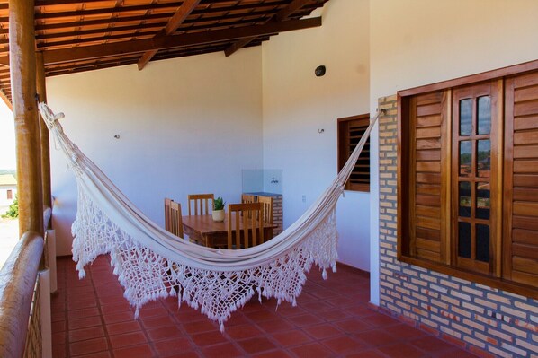 Una hamaca colgada sobre un patio con una mesa y sillas de madera, un muro de ladrillo y una ventana con contraventanas de madera.