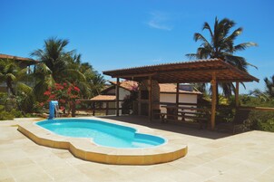 Uma piscina com borda curva, rodeada por palmeiras e um espaço coberto com área de estar.