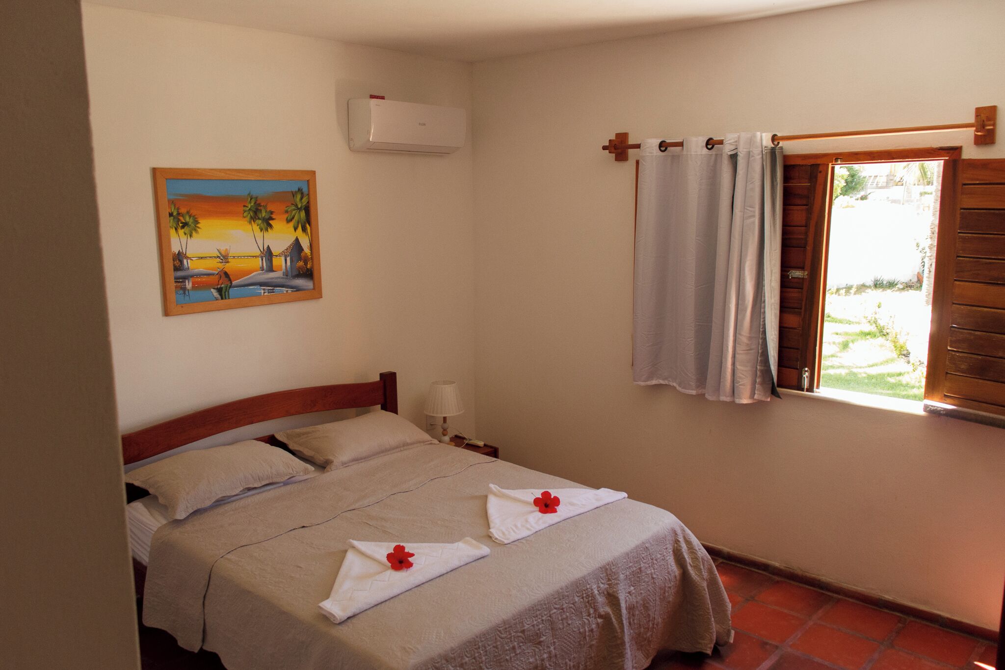 A bedroom with a bed, a painting on the wall, an air conditioner, a window with curtains, and a view of the outdoors.