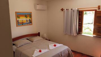 A bedroom with a bed, a painting on the wall, an air conditioner, a window with curtains, and a view of the outdoors.