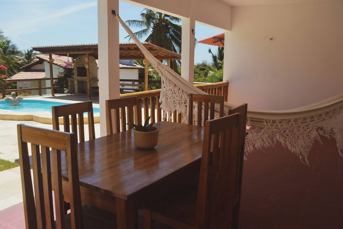 A wooden dining table with chairs, a hammock, and a view of a swimming pool and palm trees.