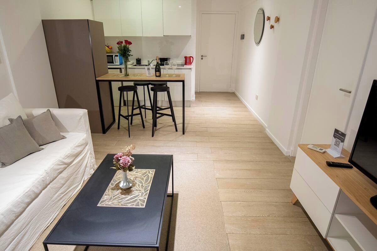 A modern living room with a sofa, a coffee table with a vase of flowers, a dining area with a table and chairs, a kitchen in the background, and a TV on a stand.
