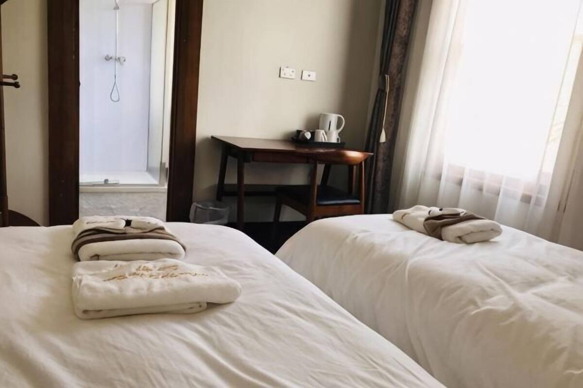 A hotel room with two beds, a small table with a kettle, and a shower area visible through an open door.