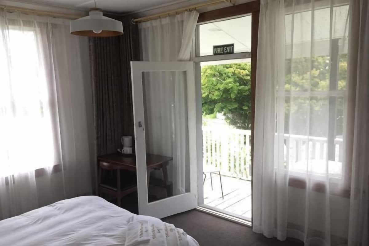 A bedroom with a bed, a desk, a chair, a window with a view of the outdoors, and a door marked 'Fire Exit'.