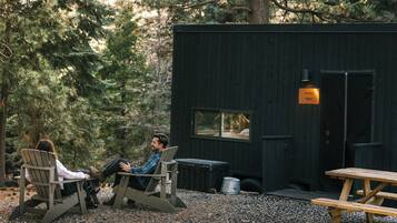 Eine Hütte mit zwei Adirondack-Stühlen davor, einem Picknicktisch und einer kleinen, schwarzen Struktur mit Fenster und Schild.