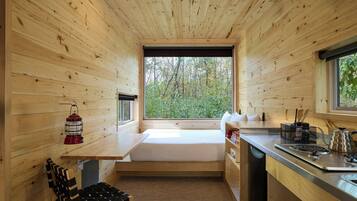 Una cabaña compacta con interior de madera, un banco empotrado, una mesa pequeña, una ventana con vista a árboles y una kitchenette con estufa y fregadero.