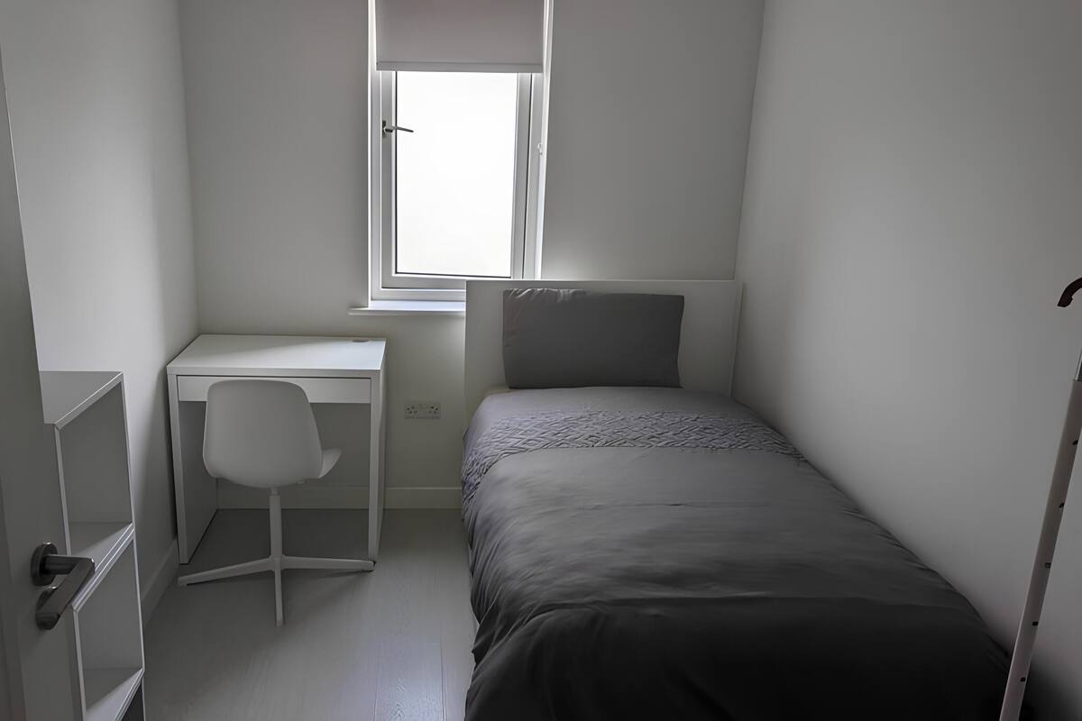 A small, sparsely furnished room with a single bed, a desk, and a chair.