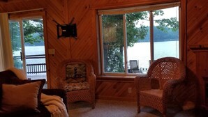 Een gezellig chalet met een groot raam dat uitzicht biedt op een meer, rieten stoelen, een open haard en een aan de muur bevestigde televisie.