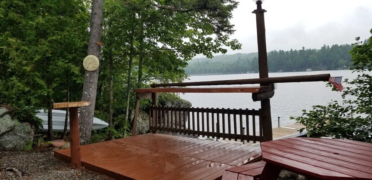 A wooden deck with a bench and a table, overlooking a lake with trees in the background.